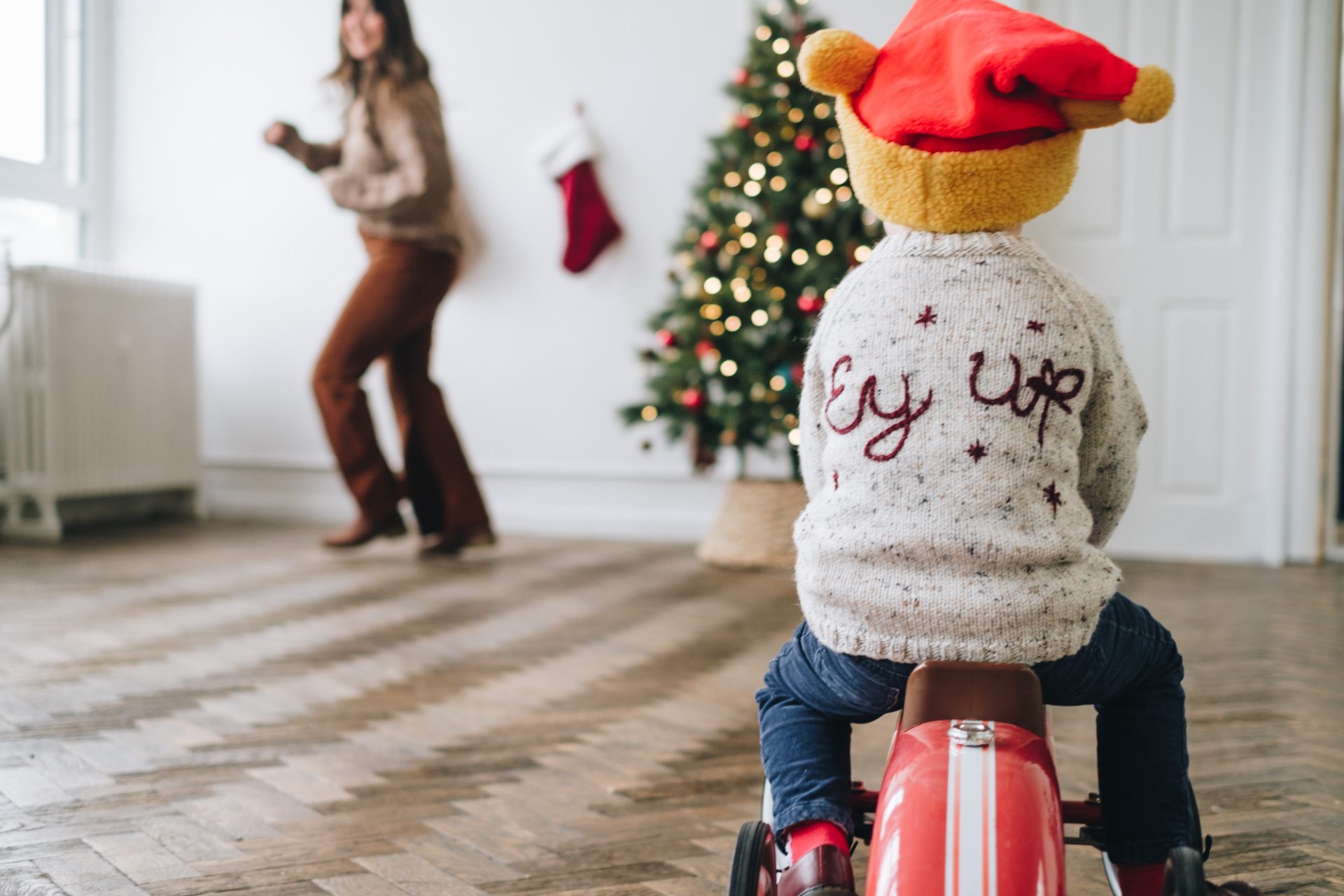 Boy on red car chasing his mum with a Christmas tree in the background