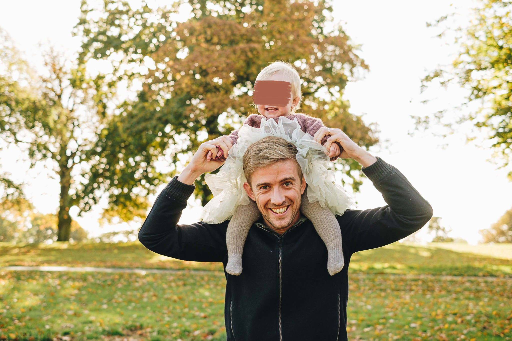 Dad holds his daughter on his shoulders in the park. Trees have yellow leaves and golden sunlight peeks through